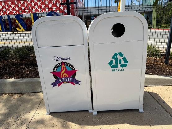 Image of two trash cans at Disney's All-Star Music Resort. The left bin features a colorful design with a musical theme and the resort's branding, while the right bin is marked for recycling with a green recycle symbol.