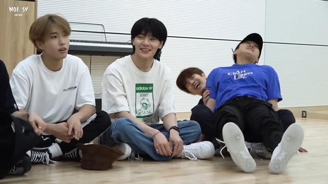 Han, I.N and Seungmin sitting on the floor of a dance practice room. Seungmin is laughing and collapsing backwards, because Changbin is sitting on his lap and leaning all his weight back agains Seungmin.
