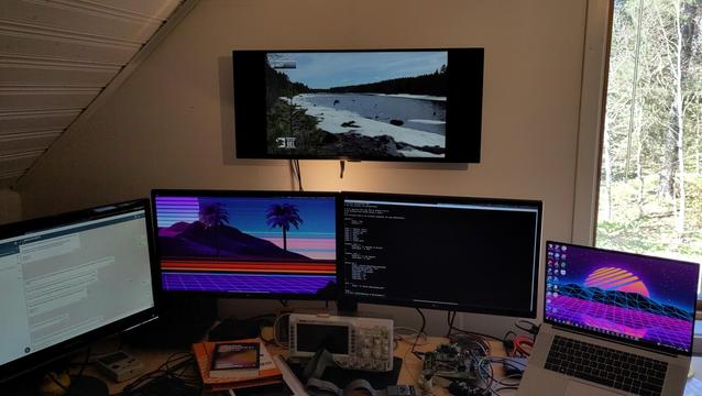 A desk with four computer screens, an oscilloscope and a fifth wide screen is mounted on the wall above the other screens and shows nature images.
