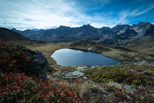 Mountain landscape with a clear blue lake, foreground of colorful foliage, and rugged peaks against a cloudy sky.