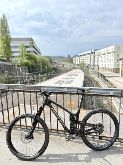 Ein schwarzes Mountainbike ist auf einem Geländer mit Blick auf die Bahngleise und Gebäude positioniert. Der Himmel ist teilweise bewölkt mit sichtbarem Grün, wodurch eine städtische Landschaft entsteht.