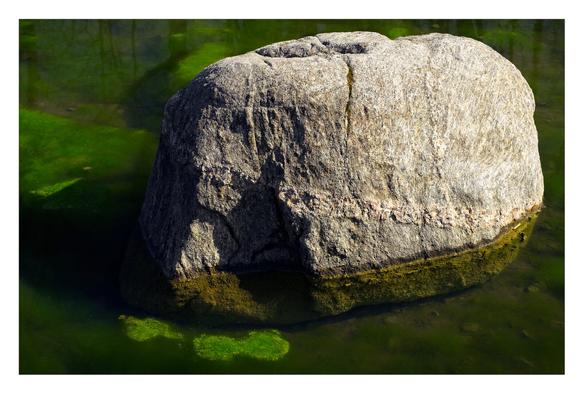 A large rock lit on an angle by the sun half emerged in green water.