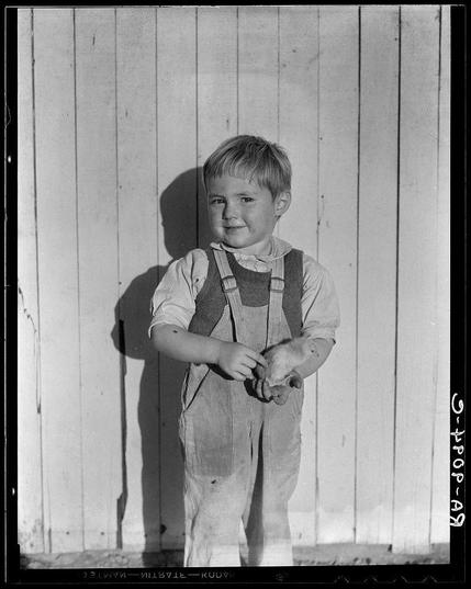 The image is a black and white photograph of a young boy standing against a plain, vertical striped background that appears to be wooden panels. The child has short hair, fair complexion, and is wearing overalls with suspenders fastened by buttons at the shoulders. He's also sporting rolled-up sleeves on his shirt which seems to have some form of pattern or embroidery around the collar area.

The boy looks directly towards the camera with a slight smile while holding what appears to be small toy animals in both hands, one held between thumb and forefinger over the other hand. His stance is upright but casual, suggesting he may not be overly comfortable posing for the picture.

There's no significant movement or action depicted apart from the boy handling his toys; otherwise, it looks like a still moment captured with focus on him against an unadorned backdrop that gives little context beyond rural simplicity and perhaps suggests work-related activity given his attire.
