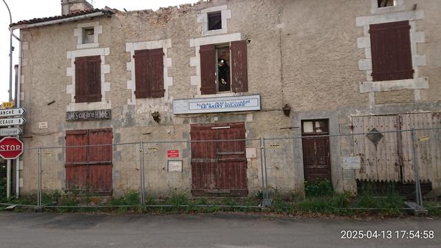 Grande bâtisse assez ancienne avec 2 pancartes : "Dames coiffure Hommes" et "Bar Auberge Le Saint-Hilaire". Le bâtiment est abandonné et le toit s'est partiellement effondré. Il est lêché par le goudron de la route...