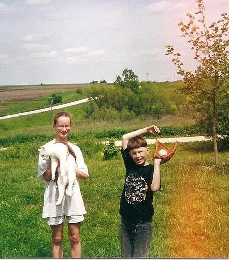 From May of 2000. An old snapshot on film.  On the right is the young sycamore tree (6 years old).  Two youngsters are in the foreground.  A teenage girl (neighbor), dressed in white, holds a large, drooping white cat (Dale the cat).  A younger boy (son) has his arms above his head.  On his left hand is a baseball glove with a baseball in the palm.