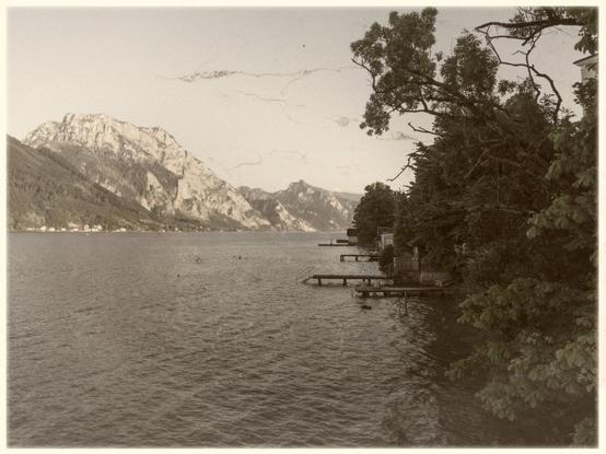 Sepia-toned photograph capturing the serene beauty of a lake framed by majestic mountains, with a tree-lined shore featuring small docks extending into the calm water.