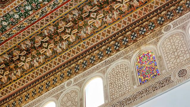 A low-angle shot showcases the intricately decorated ceiling and upper walls of Bahia Palace in Marrakech, Morocco. The ceiling features colorful, repeating geometric and floral patterns in shades of brown, green, red, and blue. The white walls have arched windows and detailed geometric carvings. A small stained-glass window with vibrant red, blue, yellow, and green pieces is visible.