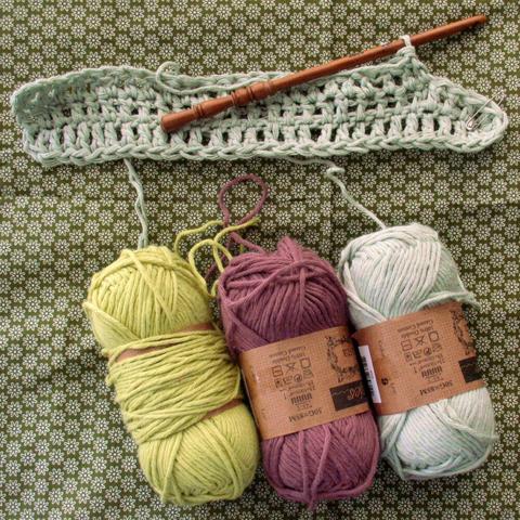 A few row of crochet stitches with a wooden crochet hook and 3 balls of yarn in lime, grey-burgundy, and light grey-green colours on a grass green fabric background with flower pattern.