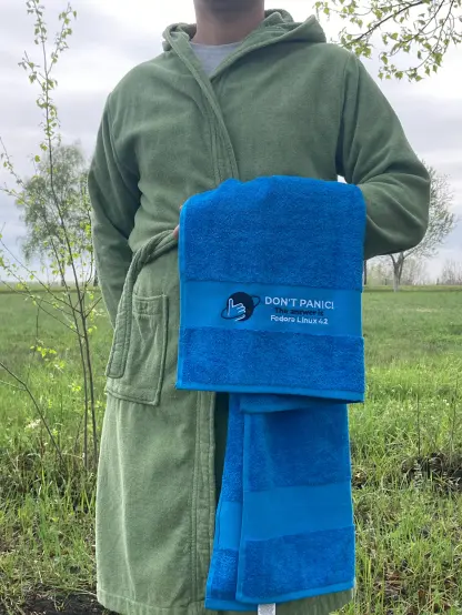 A man stands in a field. The photo cuts off his head, so the focus is on the green bathrobe he is holding... and more importantly, the towel, which is bright blue and reads

DON'T PANIC
The answer is
Fedora Linux 42

There is also a logo with a planet and a thumbs-up hand, hitchhiker style