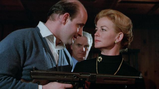 The image depicts a tense scene with three individuals indoors. On the left, a man in a blue cardigan and white shirt is looking intently at a woman to his right, while holding a submachine gun. The woman, wearing a black turtleneck and a gold brooch, gazes back at him with a serious expression. Behind them, a third man, dressed in a blue shirt and tie, observes the interaction with a stern look. The background is dimly lit, emphasizing the figures in the foreground.