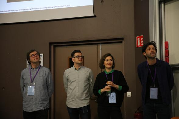 From left to right: Radim Peško, Kai Bernau, Alice Savoie, and Roland Früh standing in the back of a room with part of a conference presentation shown on a screen above their heads.