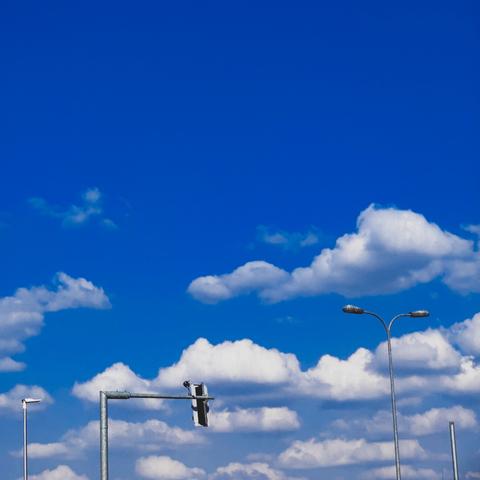 Blue skies full of small puffy white clouds, hazy with warm weather. Foreground dotted with some street lights, a traffic light, and a pole without a sign on it. Image by wannabekingpin aka vakaris