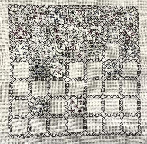 La photo représente une broderie en blackwork. Le motif se compose de 49 cases reliées par un entrelacs noir d’inspiration celtique. 25 cases ont été brodées en alternant vert et bleu et vert et violet. Elles renferment chacune un motif floral différent.