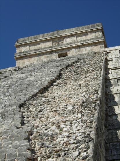 Color photo looking up at a steep staircase which ascends one of the sides of a large stone pyramid. The steps on the right side of the staircase are missing, exposing the rubble fill beneath.