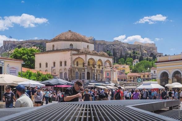 A vibrant street scene in Athens, Greece, showing a bustling crowd in Monastiraki Square with the historic Tzistarakis Mosque in the centre, colourful umbrellas shading outdoor cafes, and the Acropolis rising majestically in the background under a bright blue sky. Visible shop sign reads "GREEK SOUVENIRS".
