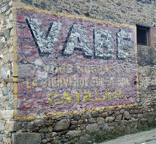 Fading painted sign on a stone-walled building. Lettering is set on a red background with a yellow border. The top half is filled with the word Vabe in white with a black shade, while the bottom has smaller yellow copy, ending with the word 'Catalan'.
