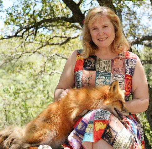 Color photo of writer Randee Dawn, a fair-skinned woman with large blue eyes and blonde hair worn long and curled, with bangs. She wears a sleeveless dress printed with vintage book covers and sits in front of a blurred background of leaves. She smiles tenderly at the camera, perhaps because she is holding a red fox on her lap. The fox is stretched out, eyes closed, looking like it’s enjoying neck scritches.