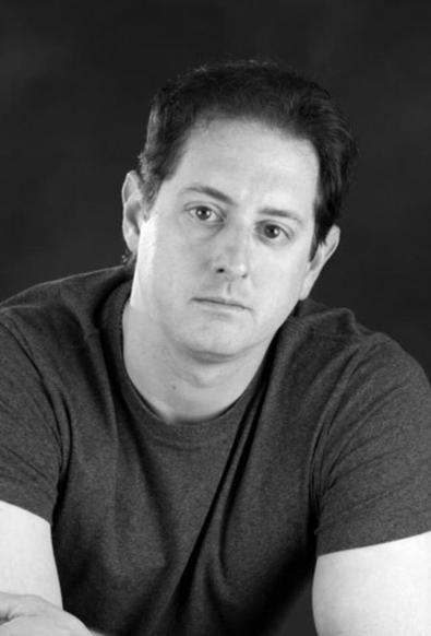 Black-and-white headshot of writer Brian Trent, a fair-skinned man with full, dark hair worn short and brushed back and large, dark eyes. He wears a short-sleeved marled tee or fine-knit medium-toned sweater and sits in front of a neutral backdrop. He leans forward slightly, head tilted a little to one side, and regards the camera with a serious expression.