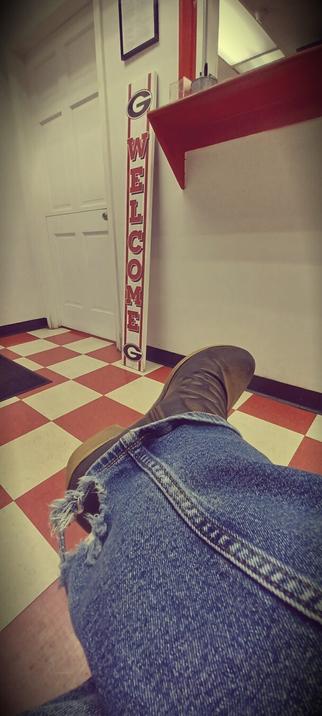 A red &amp; white checkered floor sets the backdrop for a denim clad leg ending in a brown boot. The right leg is crossed over the left. At the ankle, the jeans have a hole in them eaten away by stepping on the hem of the pants.   The photo is taken in a hot-dog pick-up stand. A red and white welcome sign is propped against the wall next to the check out window.