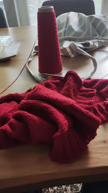 Auf einem Tisch eine fast leere Kone mit rotem gefachtem, also nicht verzwirntem Garn, eine Mischung aus Wolle, Kaschmir und Seide. Davor ein angefangenes Strickstück