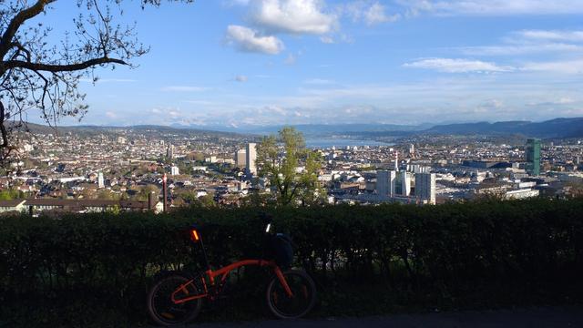 Blick von der Waid auf Zürich, den Zürisee, den Zimmerberg und in der Ferne die Alpen. Oben blauer Himmel mit ein paar weissen Wattenwolken, im Vordergrund lehnt im Schatten ein orangenes Brompton Electric G-Line an einer Hecke. Links schauen ein paar knorrige Baumäste mit jungen, kleinen Blättern ins Bild.