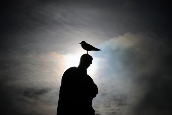 Statue des römischen Dichters Ovid in Constanta mit Möwe auf dem Kopf im Gegenlicht. Die Statue und die Möwe sind schwarz. Die Sonne hinter den Wolken steht hinter dem Kopf der Statue. Die Wolken schimmern in mystischen Farben.