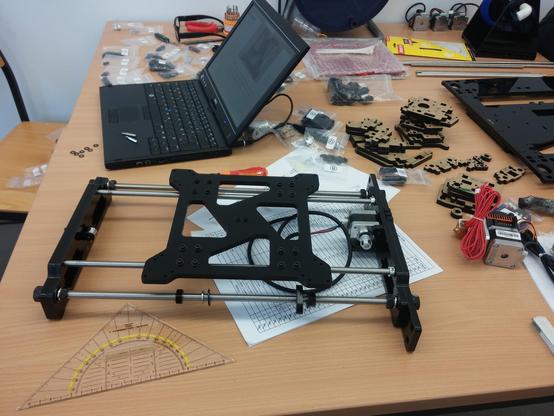 Ein Tisch mit Werkzeug, zahlreichen schwarzen Acrylteilen, nummerierten Tütchen mit Schrauben, dem bereits zusammengebauten Unterteil eines 3D-Druckers, einigen Schrittmotoren und einem Laptop.