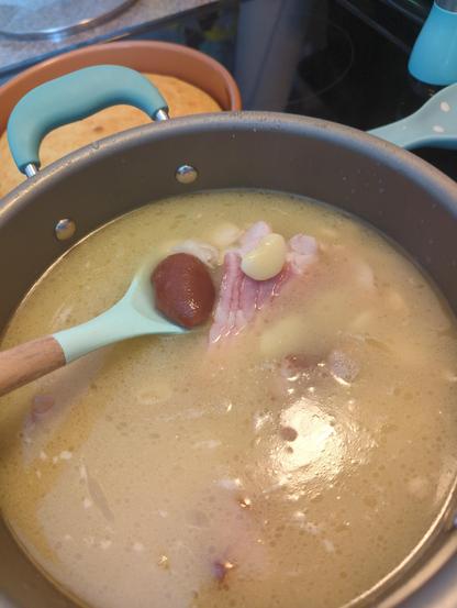 The image shows a pot of soup on a stovetop. The soup has a creamy, light brown color and contains visible ingredients such as a piece of pinkish meat, possibly ham, a whole red tomato, and a clove of garlic. A light blue spoon with a wooden handle is partially submerged in the soup, with the spoon's bowl holding the tomato. The pot has a gray exterior with a blue handle. In the background, there is a brown baking dish with a round, golden-brown item, likely bread, and a blue pepper grinder. The stovetop is black, and the countertop is gray with a speckled pattern.