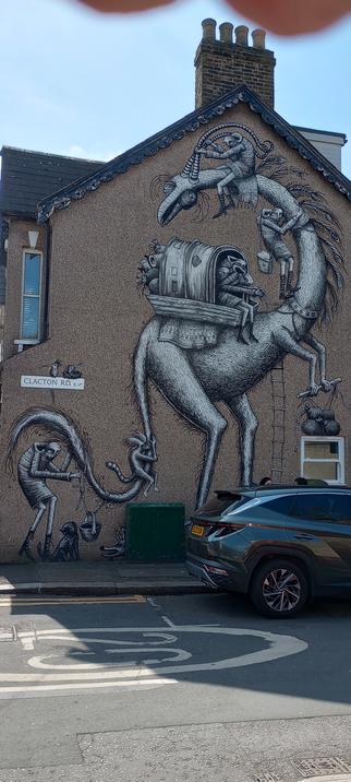 An end of a terrace with a mural. A fantastical giant two-legged creature with a long neck and long tail is shown, with a shelter on its back and various figures climbing on it.
