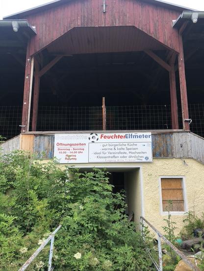 Rückseite einer Holztribüne auf Mauerwerk. Über dem vermeintlichen, dicht verwachsenen Eingang ein Werbeschild für die Gaststätte "Feuchter Elfmeter".