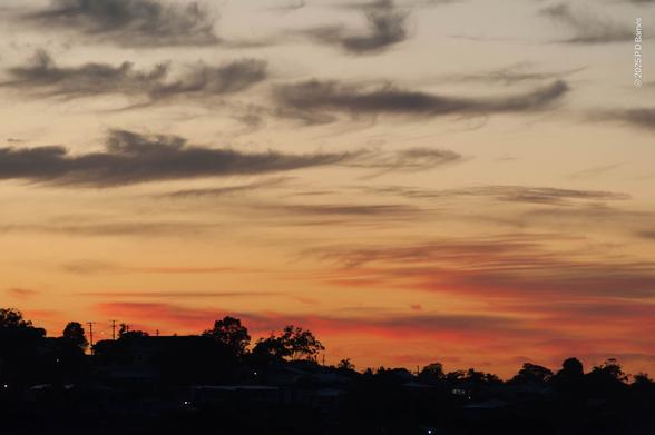 A pitch-black suburban ridgeline runs across the bottom of the frame, punctuated by a few pinpricks of light. Trees, houses and utility poles are clearly silhouetted against a deep orange-pink dawn sky. Overhead is roughly half sky and half cloud, the clouds being wind-blown, elongated and wispy. Those near the horizon are lit up pink and orange, while those higher in the sky are mid-grey.