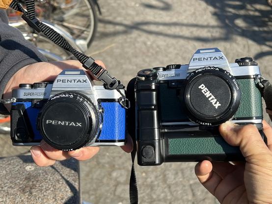 A photo of two Pentax super programs held side by side. One has blue leather, one has green leather. The green one has a big grip (the Pentax motor drive A).