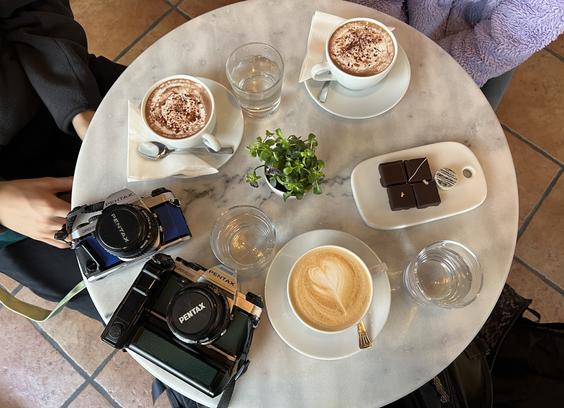 Overhead shot of a fancy table with the two cameras, a cappuccino, 2x hot chocolates, and a plate of chocolates.