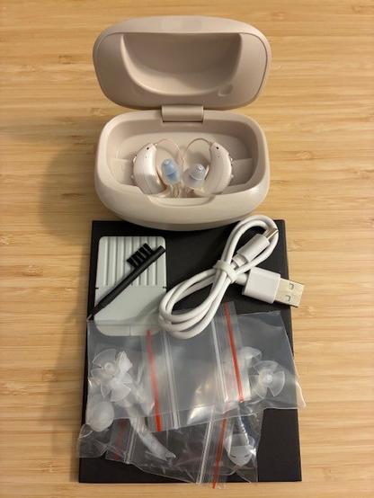 Desktop photo of low cost over the counter hearing aid set with supplied accessories, including charging case/cable, cleaning brush, replacement wax guard/tips, and silicone ear tips.
