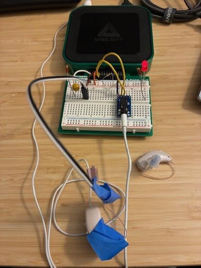 Desktop photo showing crude test setup with AD3 function generator source signal output to ear bud transducer (held near HA microphones) and in-the-canal "receiver" blue-taped next to a microphone for input to the AD3 scope input connection.