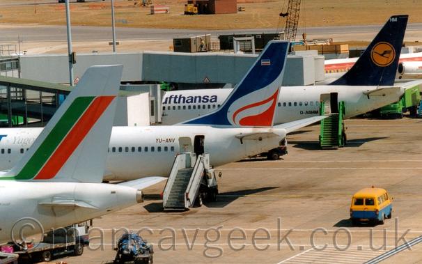 A line-up of medium-range airliners parked by a terminal building, all facing to the left, with their tails displayed prominently.
Nearest the camera, on the left, is the white rear fuselage and tail of a jet airliner, with a diagonla green and red stripe on the tail.
Next is a white jet airliner, with a blue and red tail with a white flame in the middle, and a set of white airstairs by the open rear door.
At the rear is a white jet airliner with blue "Lufthansa" titles on the upper forward fuselage, and a dark blue tail with a yellow circle containing a blue flying bird, with a green set of airstairs by the open rear door.
A blue and yellow van is driving along the concrete apron  on the right of the frame.