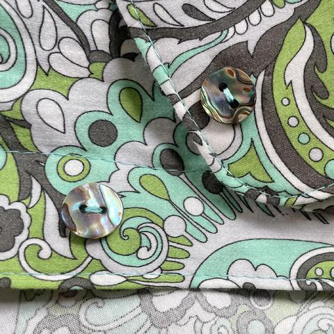 Closeup of two two-hole shirt buttons made of abalone shell that have been attached to the body of a paisley floral print shirt. Both the buttons and shirt have soft green, cyan, charcoal, and white tones. The buttons are have a pearlescent shine.
