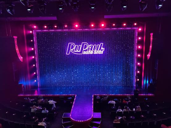 A stage set for "RuPaul's Drag Race," featuring a colorful, illuminated backdrop with a large logo. The seating area is filled with audience members, and the stage is lined with bright lights.