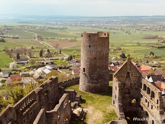Die Perspektive eröffnet einen beeindruckenden Blick auf den mächtigen runden Bergfried, das Herzstück der Burg Münzenberg. Die umliegende Landschaft der Wetterau breitet sich wie ein Teppich aus Feldern, Bäumen und kleinen Dörfern aus. Die Weite der Landschaft steht im Kontrast zur alten Steinstruktur und verleiht dem Bild etwas Erhabenes. Die Szenerie wirkt friedlich und zeitlos - als würde die Burg seit Jahrhunderten über das Land wachen.