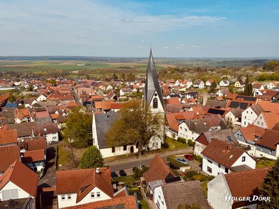 Ein stimmungsvolles Bild des Städtchens mit der markanten Kirche im Zentrum. Die spitz zulaufende Kirchturmspitze ragt stolz empor, umgeben von einem Meer aus roten Dächern. Die Umgebung blüht auf – es ist Frühling, sichtbar an den blühenden Bäumen. Das Bild vermittelt Geborgenheit, Heimatgefühl und eine gewisse heitere Ruhe.