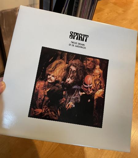 A hand holds up the album "Twelve Dreams of Dr. Sardonicus: a rectangular, very psychedelic, distorted photo of band members and some fiendish looking, face-painted, bulbous-baldheaded fellow at center with very wide white borders.