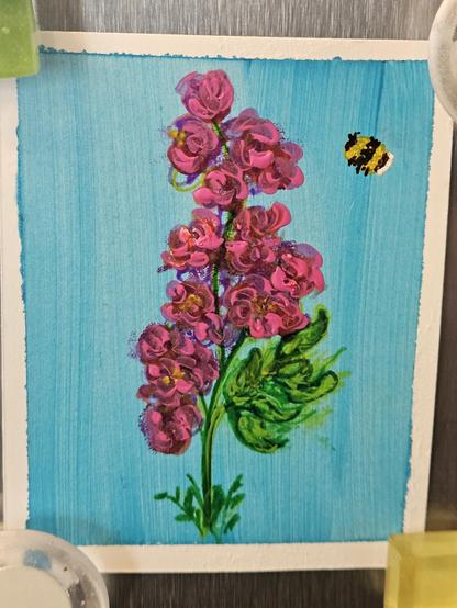 A tiny painting of some purple flowers. There is a light blue background, and a fat little yellow bumble bee about to alight.

The painting is held to a stainless steel fridge with a magnet I'm each corner.