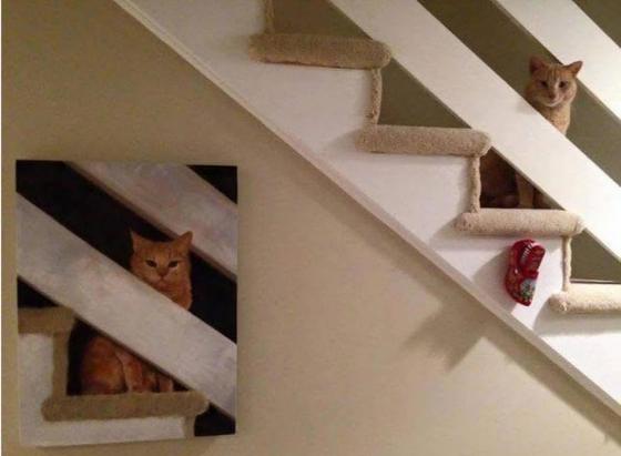 An orange tabby looks through the railing on the stairs.. Below on the wall is the painting, the exact copy of the cat at the railing.