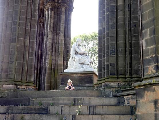 Una gran escultura de un hombre sentado, de mármol blanco, sobre un pedestal, entre enormes columnas (arcos) de piedra negruzca. En el escalón superior de la escalera que sube hasta la parte del monumento donde se encuentra la estatua, hay una niña sentada con las piernas cruzadas y la cabeza apoyada en las manos.