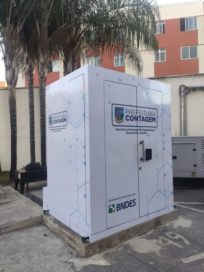 A imagem mostra um container branco com a inscrição "Prefeitura Contagem" e o logotipo da Secretaria Municipal de Planejamento, Orçamento e Gestão. O container está localizado em um espaço externo, com uma palmeira visível à esquerda e um edifício com paredes em tons de laranja e branco ao fundo. O container tem um design moderno, com linhas geométricas azuis que lembram circuitos eletrônicos. Na parte inferior, há a inscrição "Equipamento financiado pelo BNDES" com o logotipo do banco. A porta do container tem um fecho e uma maçaneta. O ambiente parece ser um espaço público, possivelmente um estacionamento, com uma calçada e uma área de concreto ao redor do container.
