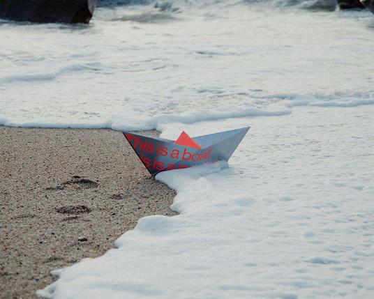 A paper boat with red text saying "This is a boat" is partially submerged in sea foam at the edge of a sandy shore.