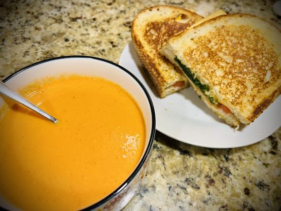 Bright orange creamy smooth soup in a bowl next to a grilled sandwich cut in two on a plate. The green spinach, red tomato, and meaty vegan cheese are protruding from the sliced side.
