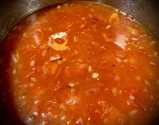 Looking into a steel pot, holding bright red diced tomatoes and water and onions.