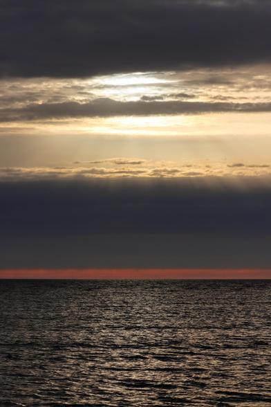 Sonnenaufgang.
Über dem schwarz-grau glitzernden Meer ein schmutzig roter Horizont. Darüber dunkelgraue Wolken. Und über allem eine tiefstehende Sonne, die ihr Licht durch die Wolken schickt.