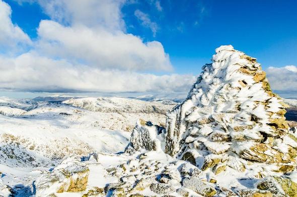 A tall cairn fills most of the right half of the frame, mostly plastered in crystals of rime-ice. Snow and more ice cover the foreground with a few exposed bits of yellow-gray rock. More rugged snowy hills roll away into the distance under a part cloudy part blue sky.